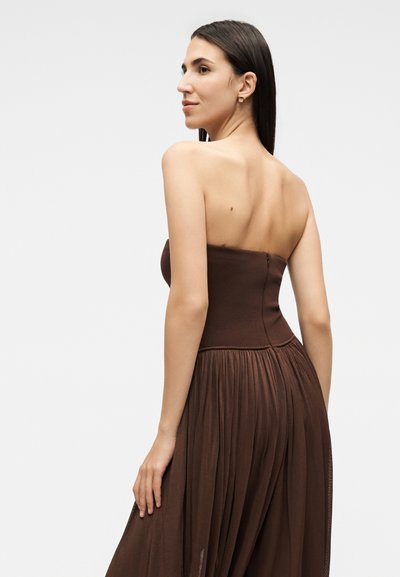 Γυναίκα με ίσια σκούρα μαλλιά, που φοράει ένα καφέ πλισέ φόρεμα χωρίς τιράντες, γυρισμένη πλάτη και κοιτάζει πάνω από τον ώμο της, με φόντο έναν απλό χώρο.