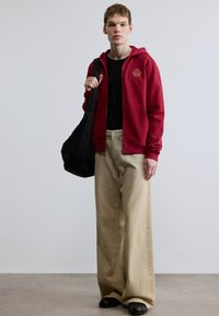 Červená mikina na zips s logom, čierne tričko, nadrozmerné béžové nohavice a čierna taška. Hladká tkanina, uvoľnený strih a minimálne detaily.