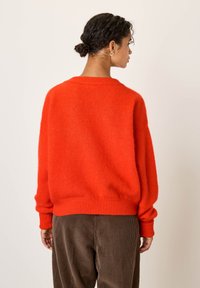 Personne aux cheveux bouclés foncés attachés en chignon bas, portant un pull orange vif en laine duveteuse et un pantalon en velours côtelé marron, de dos sur un fond uni.