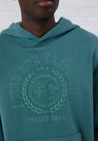 Hoodie turchese con cappuccio, caratterizzato da una grafica in rilievo di frutta circondata da alloro e il testo "Pier One Classics Health Club".