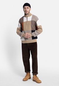 Jersey de punto con un diseño de bloques de colores en beige, marrón y blanco, con un cuello de media cremallera y patrones texturizados. Combinado con pantalones de pana marrones y botas beige.