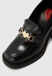 Mocassins en cuir noir avec une semelle épaisse et un matériel en forme de cœur doré portant l'inscription "LOVE MOSCHINO" sur la sangle.