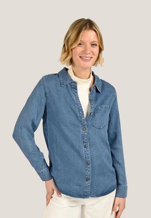 Femme souriante aux cheveux blonds courts, portant une chemise en jean boutonnée sur un top blanc, debout avec une main sur la hanche devant un fond uni.