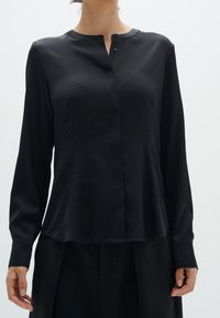 Svart långärmad blus i slät tyg, med en bandkrage, diskret knäppning och en avslappnad passform. Enkelt design utan mönster.