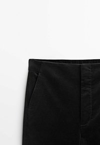 Pantalon en velours côtelé noir à la taille et sur la partie supérieure de la cuisse avec poche latérale et fermeture éclair avant sur fond blanc.