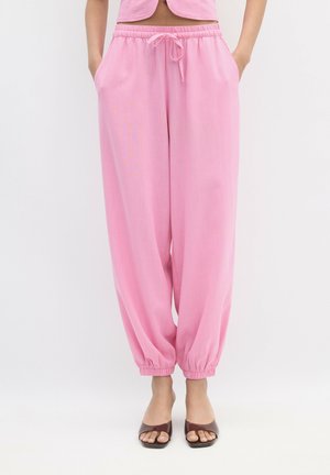 Femme portant un pantalon rose ample avec cordon de serrage et poignets élastiques, ainsi que des talons ouverts marron, debout les mains dans les poches sur fond blanc.
