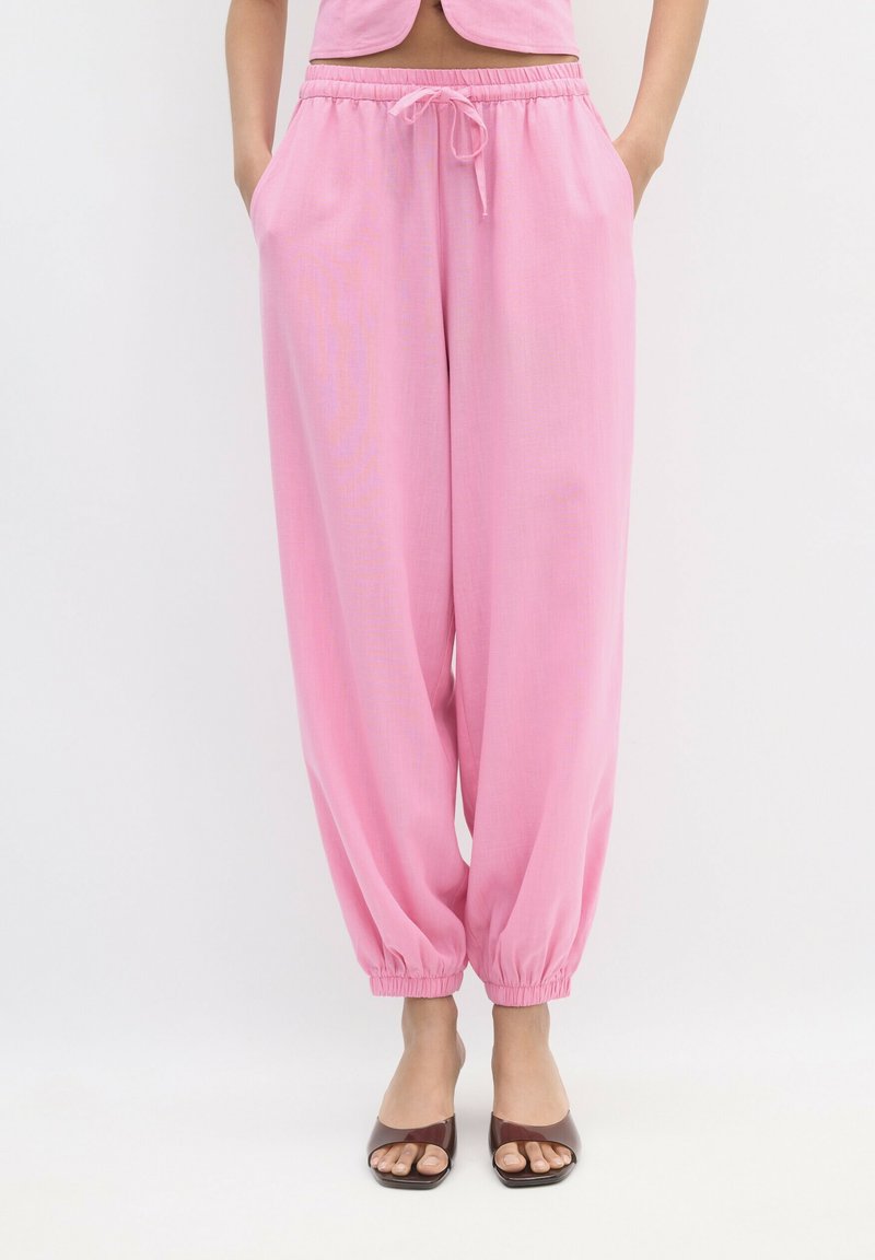 Femme portant un pantalon rose ample avec cordon de serrage et poignets élastiques, ainsi que des talons ouverts marron, debout les mains dans les poches sur fond blanc.