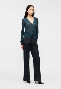 Σκούρο τιρκουάζ ελαφρύ πλεκτό ζακέτα με V-λαιμόκοψη και κλείσιμο με κουμπιά, σε συνδυασμό με ψηλόμεσο σκούρο τζιν παντελόνι και μυτερές μαύρες γόβες.