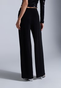 Pantalons larges noirs en tissu scintillant, présentant une texture lisse et une taille haute. La silhouette est décontractée, tombant jusqu'au sol.