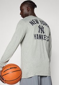 Langermet grå skjorte med "New York Yankees" trykk i marineblått på ryggen, båret av en person som holder en basketball. Atletisk passform, myk tekstur.