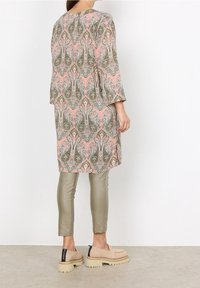 Tunique longue et fluide avec un motif paisley en rose, vert et blanc. Associée à un pantalon slim brillant de couleur vert olive et des chaussures à enfiler beige.