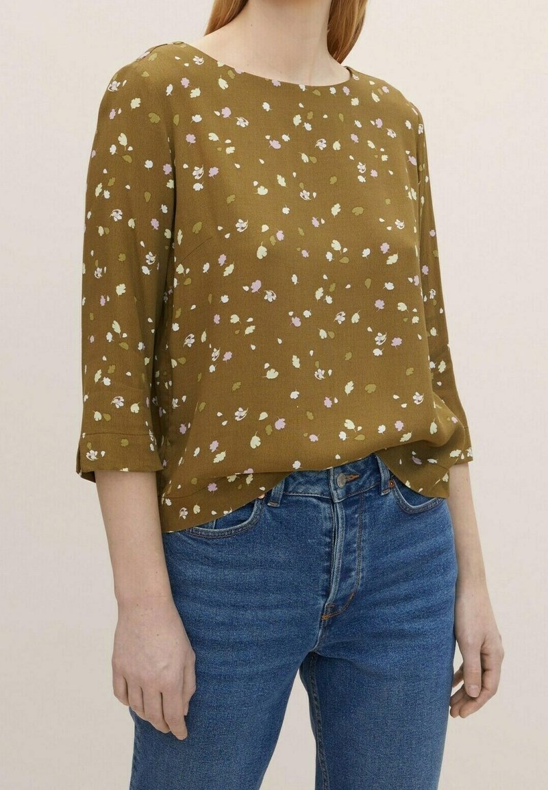 Olivgrüne Bluse mit lockerer Passform, 3/4-Ärmeln, verstreuten floralen Mustern in Rosa und Weiß, kombiniert mit blauen Jeans.
