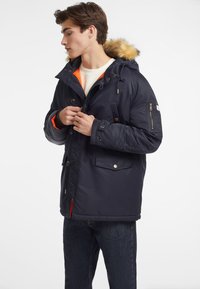 Parka azul marino con capucha forrada de piel, bolsillo con cremallera en la manga y forro naranja. Presenta botones a presión y bolsillos laterales, fabricada con material resistente al agua.