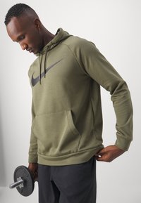 Sudadera verde oliva con el logotipo de Nike en negro, que cuenta con un bolsillo tipo canguro, capucha con cordón y puños de canalé. Pantalones cortos de entrenamiento grises.