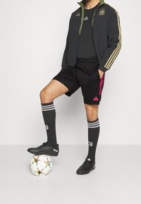 Chaqueta deportiva negra con forro verde, camiseta negra, pantalones cortos negros, calcetas negras con rayas blancas, zapatos de fútbol negros y un balón de fútbol multicolor.