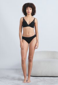 Μαύρο σετ εσωρούχων αποτελούμενο από σουτιέν και μπικίνι με λουλουδάτα σχέδια, κυματιστές άκρες και λεπτά αξεσουάρ. Το μοντέλο στέκεται σε έναν ανοιχτό γκρι φόντο.