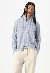 Light blue plaid button-up shirt with long sleeves, made of textured fabric. Paired with cream-colored drawstring pants.