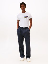 Witte T-shirt met korte mouwen en een logo in meerdere kleuren, gecombineerd met donkerblauwe, ruimvallende, gestreepte broeken en witte sneakers, staand tegen een lichte achtergrond.