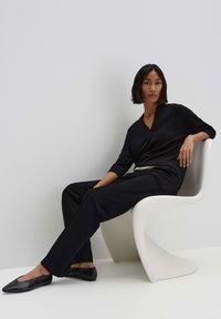 Zwarte v-hals top en losse zwarte broek met platte zwarte schoenen. Model zit op een witte sculpturale stoel, met gladde texturen en een minimalistisch ontwerp.