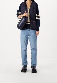 Námořnická prošívaná vesta přes světlý svetřík, doplněná světlými modrými džíny se rovnými nohavicemi a černými botami na platformě. Drží černou kabelku.