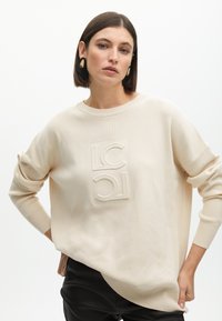 Maglione beige lavorato a maglia con scollatura rotonda, caratterizzato da un logo in rilievo sul davanti. Maniche lunghe con polsini arrotolati e orlo curvo.