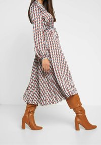 Dlouhé vzorované šaty s geometrickými tvary v červené, modré a krémové barvě. Vybavené hnědými kozačkami nad kolena a dlouhými rukávy.