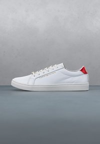 Baskets basses blanches avec lacets blancs, texte doré "Tommy Hilfiger" sur le côté, et languette de talon rouge, sur une surface et un fond gris.