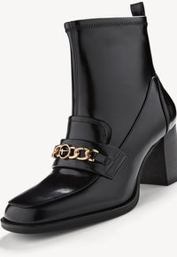 Bottine en cuir noir avec talon bloc, bout pointu et détail chaîne dorée sur le dessus.