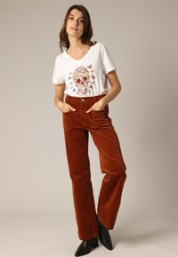 T-shirt blanc à col en V avec impression de skull et de papillon, associé à un pantalon en velours côtelé rouille taille haute avec des poches latérales et des bottes noires à talons.