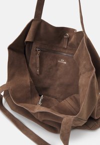 Borsa a tracolla in suede marrone con un ampio interno, dotata di una tasca con zip, dalla texture morbida e con lunghe tracolle. Dettagli di branding minimal visibili.