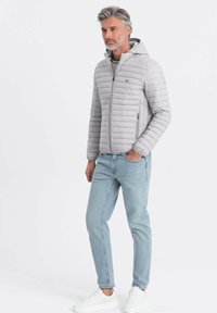 Lys grå pufferjakke med hætte, der har vandret quiltet design, sidelommer og lynlåslukning, båret med lyse jeans og hvide sneakers.