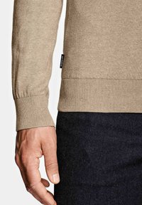Poignet et ourlet d'un pull beige en maille à manches longues avec une étiquette en tissu noir à côté de la main sur un fond de pantalon foncé.