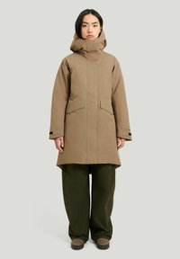 Brun vattentät parka med hög krage, stora framfickor och rak passform, tillsammans med gröna löst sittande byxor och bruna skor.