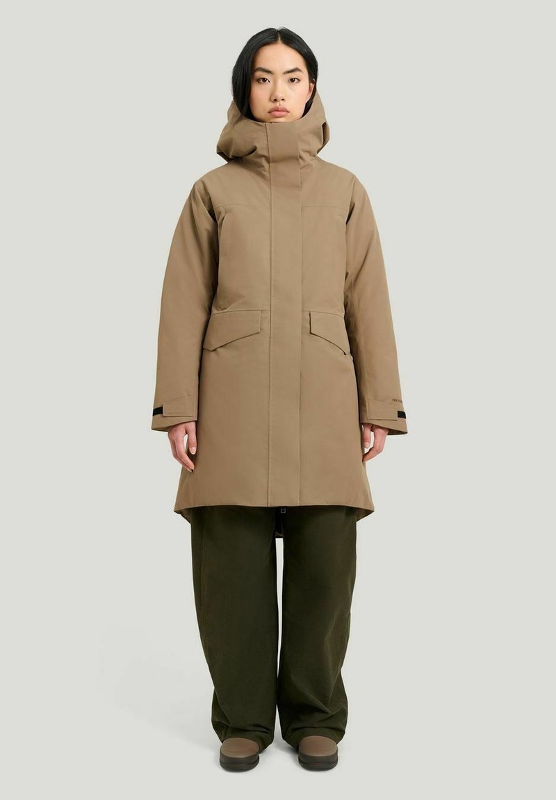 Brun vattentät parka med hög krage, stora framfickor och rak passform, tillsammans med gröna löst sittande byxor och bruna skor.