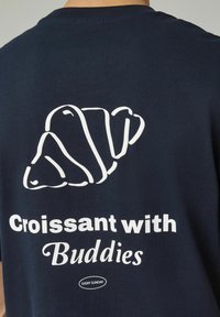 T-shirt bleu marine avec un graphisme en contour blanc d'un croissant au-dessus du texte "Croissant avec des Amis" dans une police ludique.
