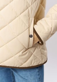 Pehmest beežist tikitud jakk pruunide ääristega, millel on klõpslukustus ja küljetasku. Sile tekstuur teemantkujundusega detailiga.