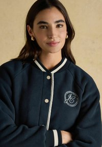 Tumesinine fliisist varsity jope, millel on valge tõmblukk, broderitud logo ja triibulised soonikud. Pehme tekstuur ja lõdvestatud sobivus.