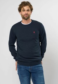 Marineblauwe gebreide trui met ronde hals, geribbelde manchetten en zoom. Heeft een klein rood logo op de linkerborst. Gecombineerd met blauwe spijkerbroek.