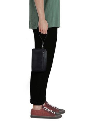 Person in green shirt and black pants holding a black zippered wristlet pouch beside their leg, wearing red canvas sneakers with gray toes.