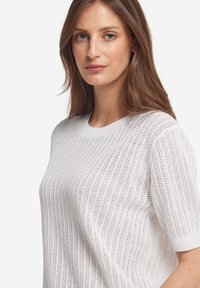Hvid strikket sweater med ribbet tekstur, korte ærmer og rund hals design. Stofmet er præget af et mønstret åbent væv.