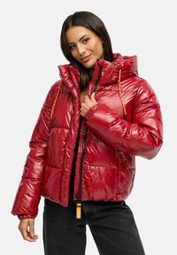 Chaqueta acolchada roja con un acabado brillante, cierre de cremallera, capucha con cordón y puños elásticos. Presenta detalles en naranja y un diseño cuadrado.