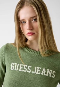 Maglione verde lavorato a maglia con colletto a coste e testo bianco "GUESS JEANS" sul davanti, dalla consistenza morbida.