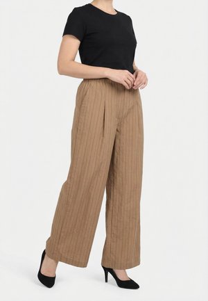 Femme portant un haut noir à manches courtes, un pantalon beige à rayures fines taille haute et jambes larges, et des chaussures noires à talons hauts, debout devant un fond blanc.