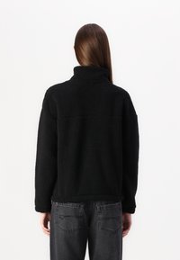 Zwarte fleecejack met een hoge kraag, gestructureerd oppervlak en een losse pasvorm, gecombineerd met donkere, recht gesneden jeans met achterzakken.