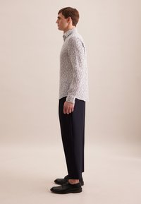 Witte lange mouwen shirt met een multicultureel bloemenpatroon, gecombineerd met zwarte op maat gemaakte broeken en zwarte schoenen, weergegeven in profielweergave.