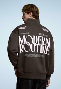 Brązowa, luźna bluza z dużym białym napisem na plecach, z wysokim kołnierzem i ściągaczami na rękawach. Gładka tkanina o swobodnym kroju.