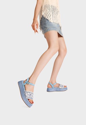 Donna che cammina con sandali platform blu con cinturini floreali, indossa un top all'uncinetto e una minigonna di jeans su uno sfondo semplice.