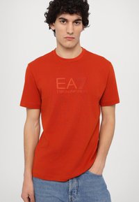 Red cotton t-shirt with a round neck and short sleeves, featuring a prominent printed logo "EA7 Emporio Armani" in the center.