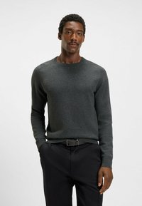 Maglione lavorato a maglia di un verde scuro, con scollo rotondo e maniche lunghe, abbinato a pantaloni neri, caratterizzati da una texture liscia e un design aderente.