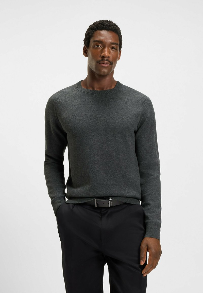 Maglione lavorato a maglia di un verde scuro, con scollo rotondo e maniche lunghe, abbinato a pantaloni neri, caratterizzati da una texture liscia e un design aderente.